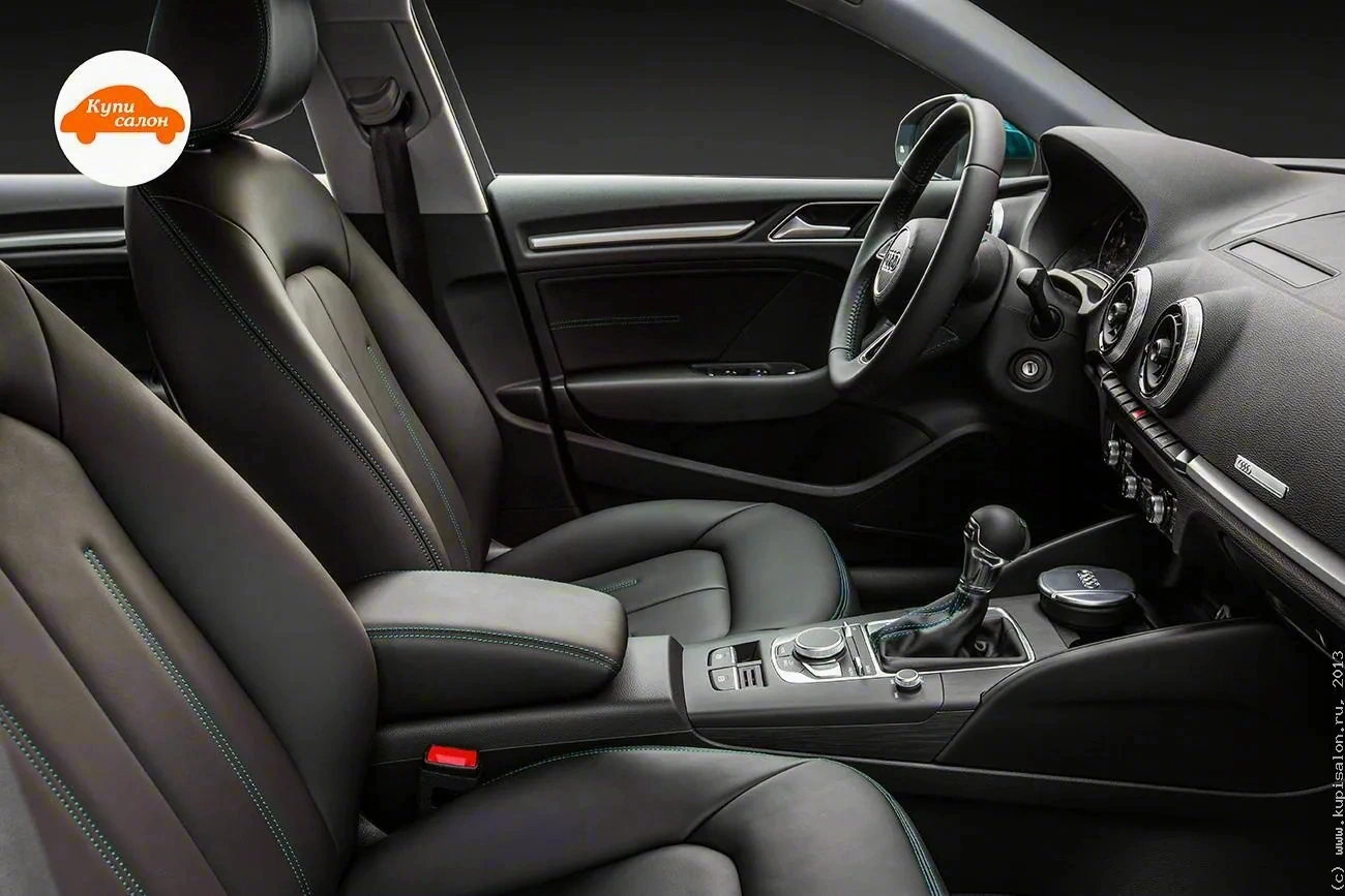 Салон Audi A3, вид со стороны переднего пассажира: передние кресла с чёрной обивкой и бирюзовой строчкой, подлокотник, рычаг коробки передач, руль и часть передней панели с круглыми дефлекторами
