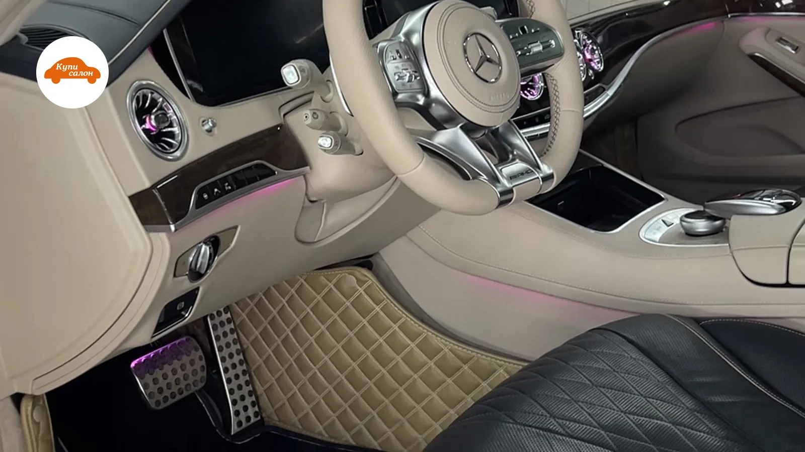 Интерьер Mercedes-Benz с бежевым рулем, черными сиденьями с ромбовидной перфорацией, бежевыми ковриками и розовой амбиентной подсветкой