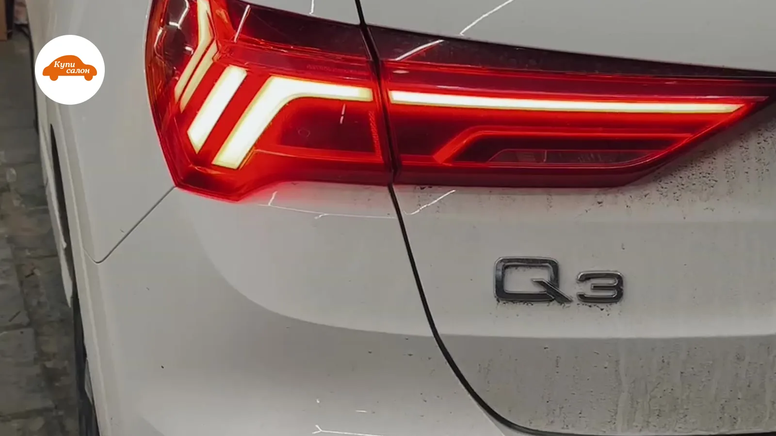 Задняя часть белого Audi Q3, видны светодиодные фонари и шильдик Q3, кузов частично грязный.