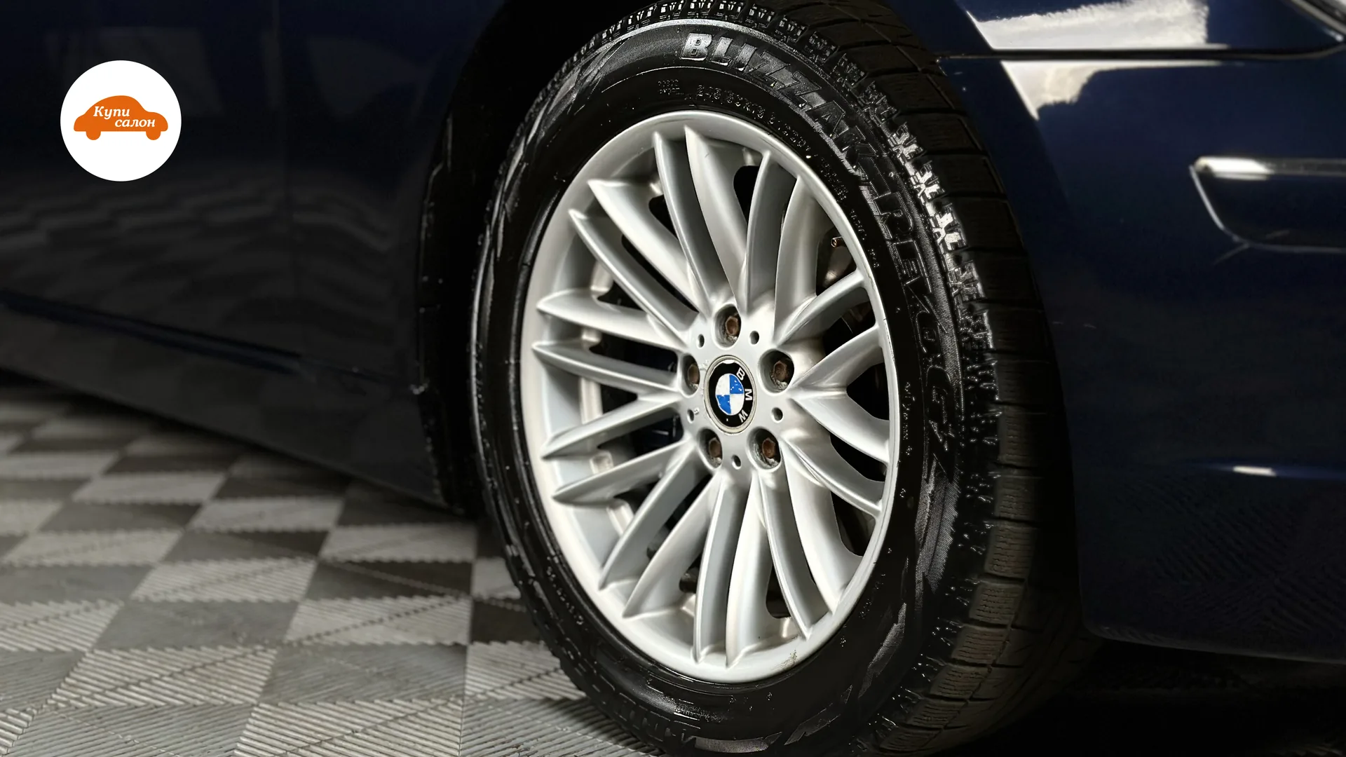 Синий автомобиль BMW, крупный план колеса и мокрой шины на клетчатом полу.