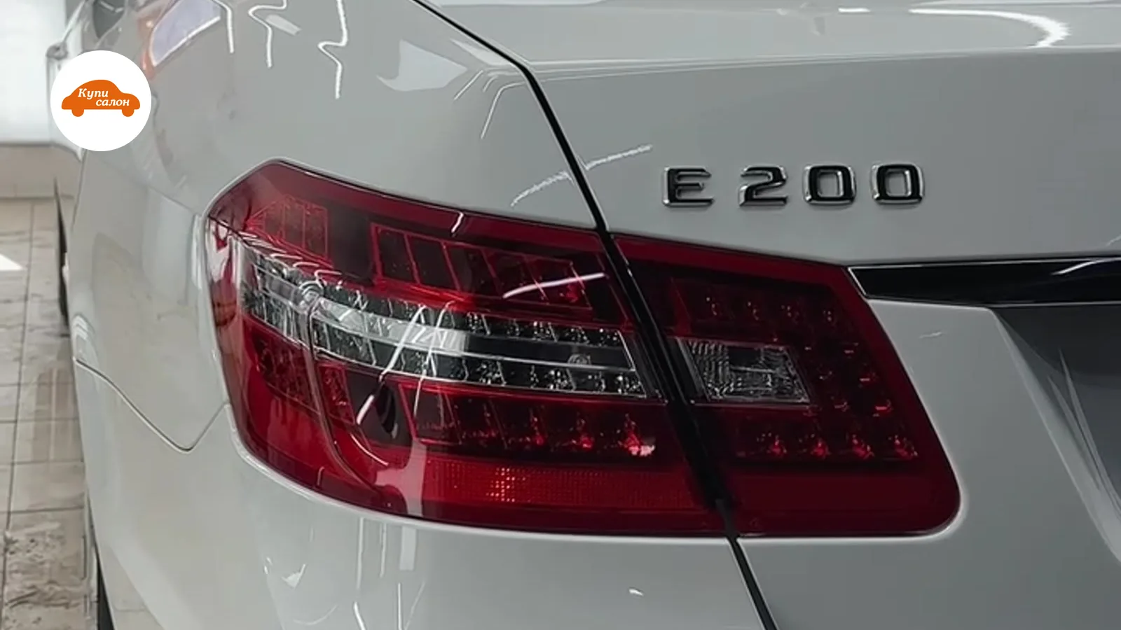 Задняя часть белого автомобиля Mercedes-Benz E 200, видны задние фонари и шильдик