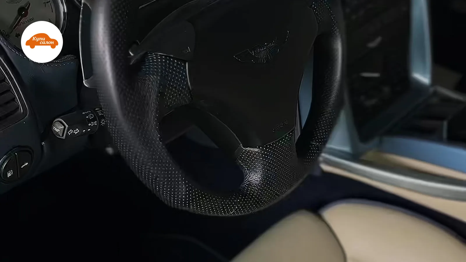 Интерьер Aston Martin с перетянутым рулем под карбон и бежевым кожаным сиденьем с ромбовидной строчкой.