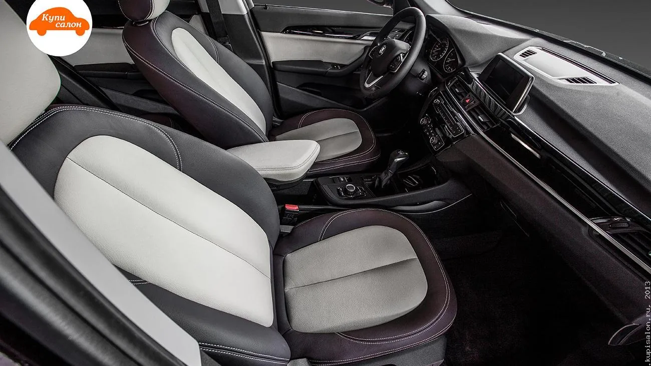 Салон BMW X1 прошлого поколения после перетяжки: передние сиденья и центральный подлокотник в тёмной и светлой отделке со светлой строчкой, консоль с рычагом селектора