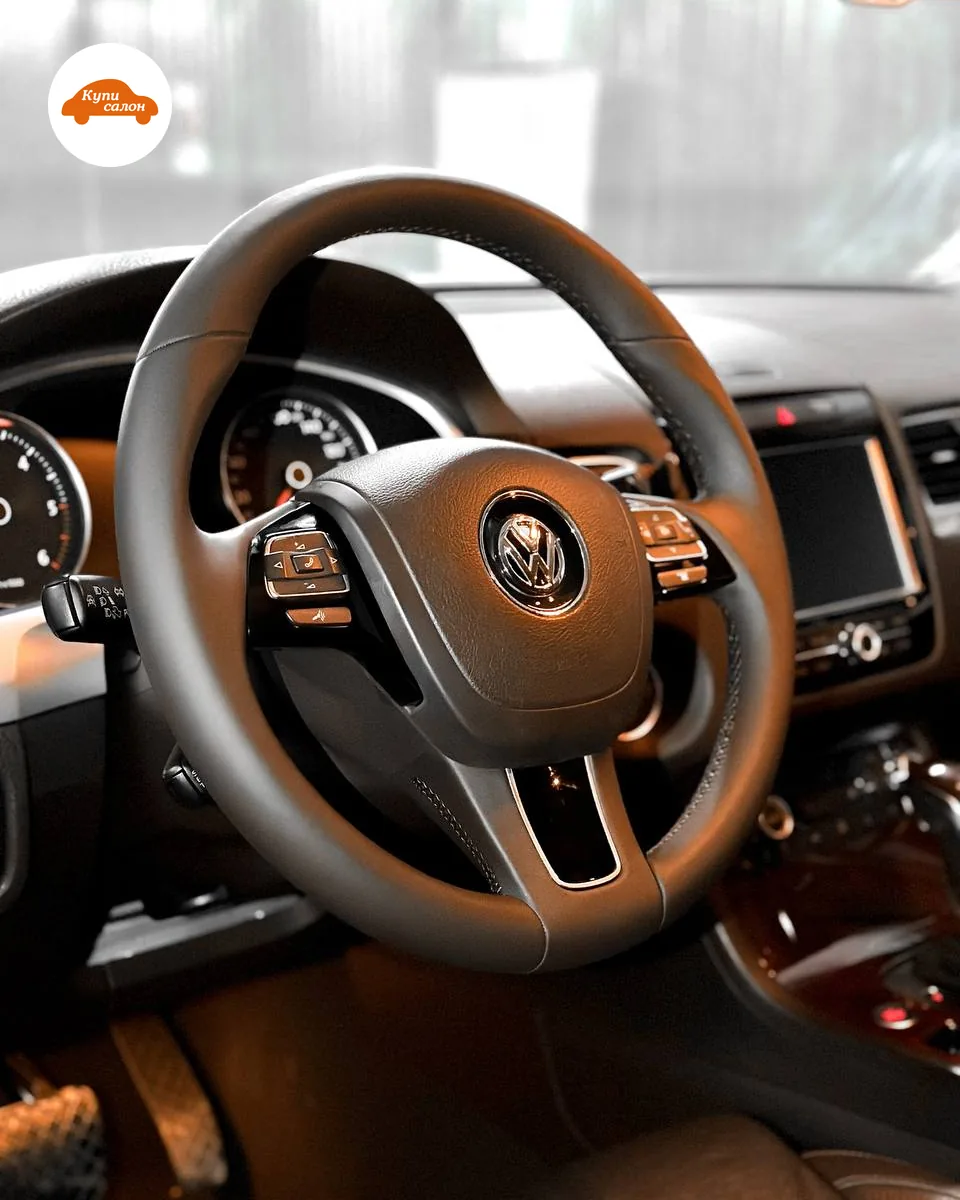 Руль Volkswagen Touareg второго поколения в чёрной коже со строчкой, вид со стороны водительской двери: блоки кнопок на спицах, за рулём приборы