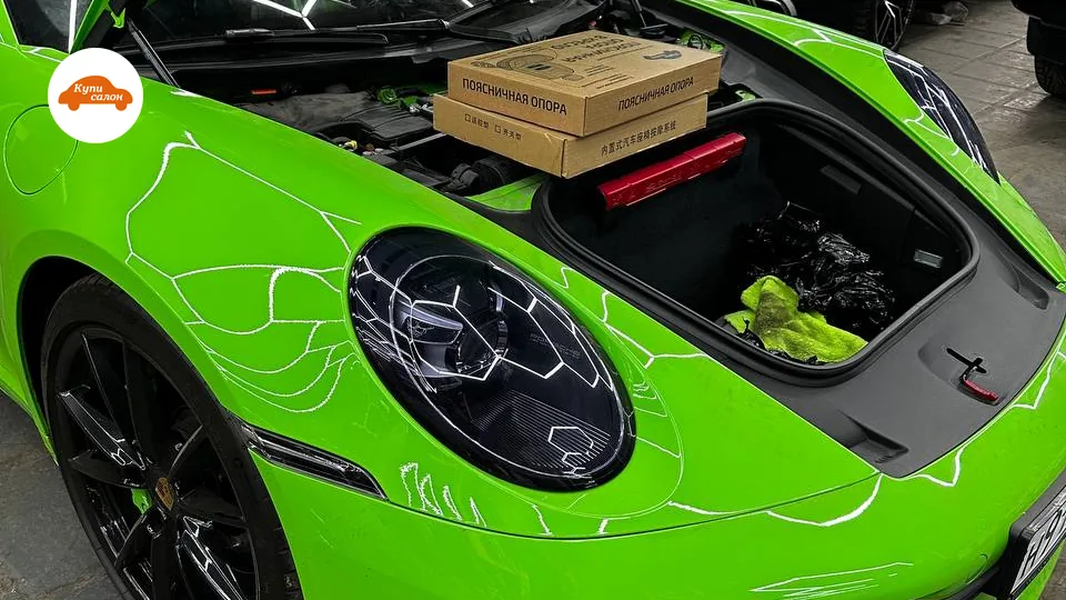 Porsche 911 (992) ярко-зеленого цвета с открытым багажником, в котором лежат коробки с поясничной опорой, кузов оклеен защитной пленкой