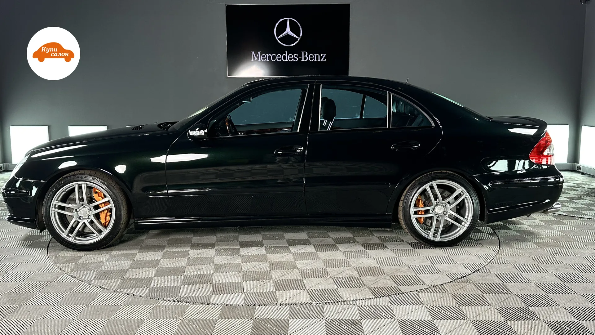 Темно-зеленый седан Mercedes-Benz E-класса, вид сбоку, на фоне стены с логотипом Mercedes-Benz