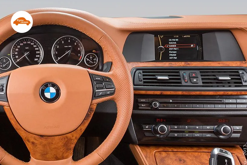 Консоль BMW 5 серии в кузове F10: экран, дефлекторы, магнитола с клавишами 1–8 и блок климата с красными цифрами температуры и двумя круглыми регуляторами; слева руль в коричневой коже