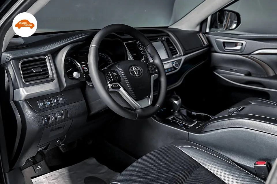 Салон Toyota Highlander третьего поколения через водительскую дверь: руль, чёрная подушка водительского сиденья с перфорацией в центре, слева от руля под дефлектором два ряда прямоугольных клавиш
