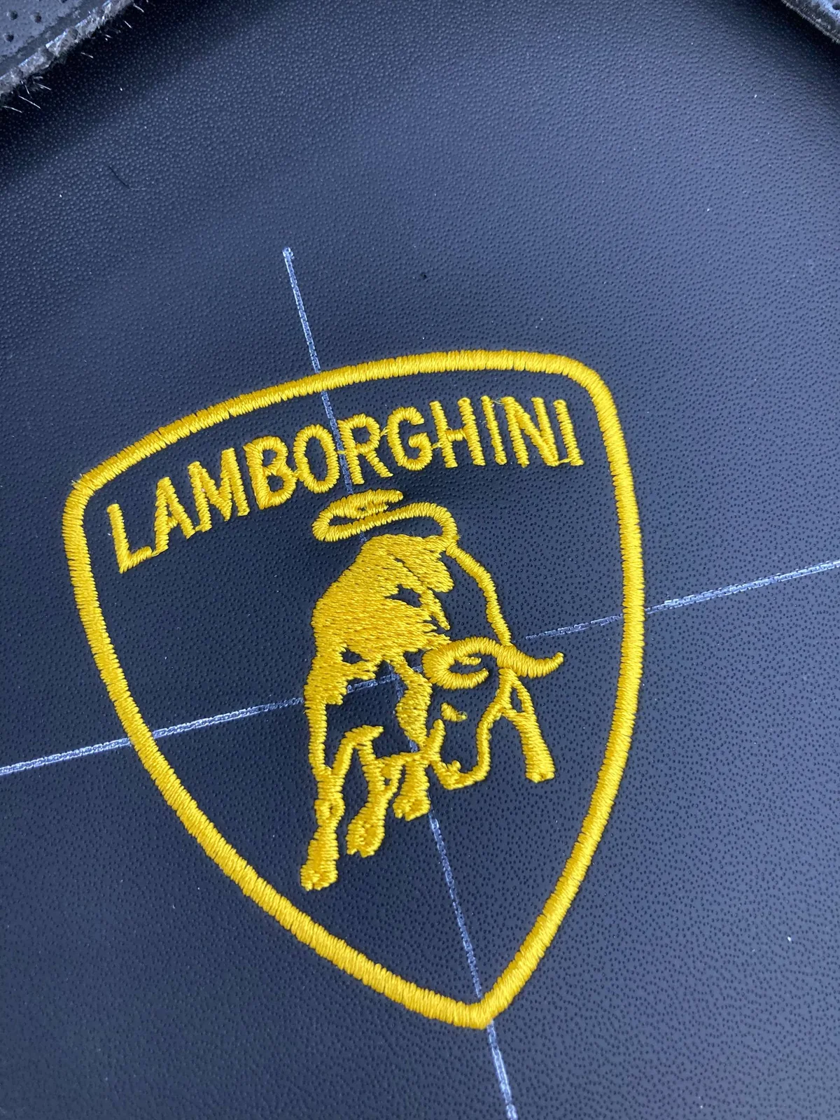 Крупный план готовой вышивки: логотип Lamborghini — щит с быком, вышитый золотой нитью на чёрной гладкой коже вставки сиденья